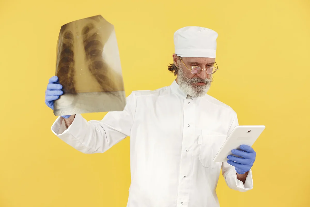 Bearded senior doctor in white coat examining an x-ray while holding a tablet ag